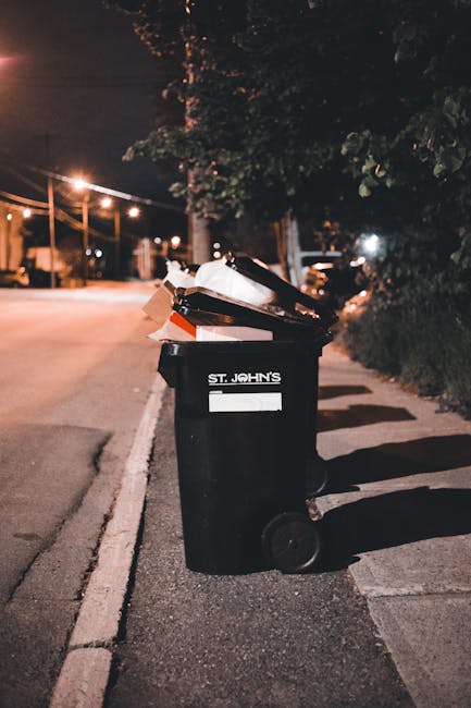 A black wheeled rubbish bin with a white label and the text 'ST. JOHN'S' printed on it is positioned on the pavement alongside a curb in an outdoor street scene at night. The bin is overflowing with various waste items, including cardboard boxes and plastic containers, some of which are leaning or resting on top. The street is illuminated by overhead streetlights, casting a warm, orange glow on the scene. In the background, there are trees with dense foliage, and blurred lights from parked vehicles and distant buildings. The setting suggests an urban environment where private waste collection or on-site rubbish disposal is being carried out, as part of an alternative waste handling service. The scene appears quiet with no visible pedestrians or moving traffic.