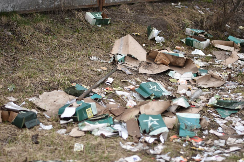 The image shows an outdoor area with a patch of grass and soil, scattered with discarded flattened cardboard boxes, some with printed logos, and torn paper waste. The cardboard appears weathered and partially crushed, with varying shades of brown and green, indicating outdoor exposure. Twisted and broken pieces of cardboard are spread across the uneven ground, with some boxes partially open, revealing white interiors. There are also loose paper scraps mixed among the cardboard debris. In the background, a metal fence is visible at the top of the frame, suggesting a boundary or boundary wall of a property or storage site. The overall scene has a dull atmosphere, with natural light highlighting the cluttered and unmanaged state of the waste. The presence of this litter indicates an instance of improper disposal or an area requiring cleanup as part of a rubbish removal service, such as those offered by Rubbish Removal Acton, to manage non-local authority waste or surplus materials from private collection efforts.
