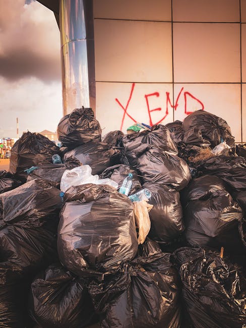 A large pile of black plastic rubbish bags piled against a modern building with tiled exterior walls, some tied tightly, with crumpled and torn sections revealing various waste contents. Several clear plastic water bottles are visible protruding from the bags, alongside other miscellaneous debris. In the background, part of a cloudy sky with soft sunlight is visible, casting light on the scene, and a graffiti message reading 'XEND' is spray-painted in red on the building's surface near the bags. The scene depicts an unmanaged rubbish accumulation likely resulting from private waste collection or on-site clearance, emphasizing the importance of professional rubbish removal services like those offered by Rubbish Removal Acton in supporting proper waste disposal. The environment appears urban, on a paved area adjacent to the building, with no other visible waste or disposal equipment present, suggesting an untreated dump site.