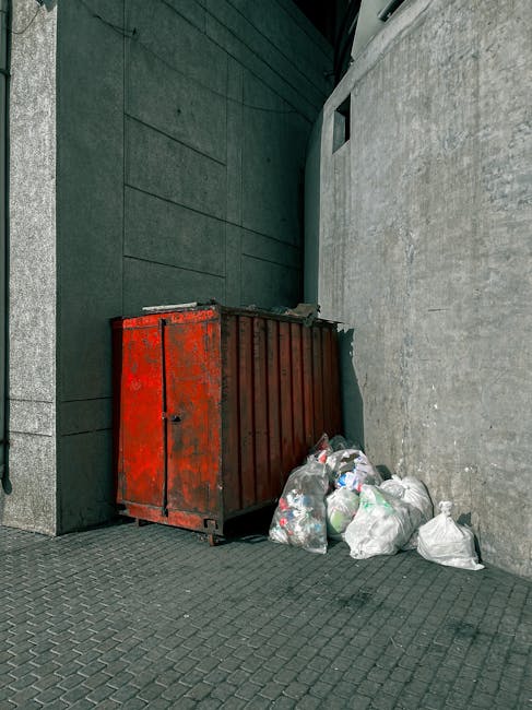 The image shows an outdoor area with a patch of grass and soil, scattered with discarded flattened cardboard boxes, some with printed logos, and torn paper waste. The cardboard appears weathered and partially crushed, with varying shades of brown and green, indicating outdoor exposure. Twisted and broken pieces of cardboard are spread across the uneven ground, with some boxes partially open, revealing white interiors. There are also loose paper scraps mixed among the cardboard debris. In the background, a metal fence is visible at the top of the frame, suggesting a boundary or boundary wall of a property or storage site. The overall scene has a dull atmosphere, with natural light highlighting the cluttered and unmanaged state of the waste. The presence of this litter indicates an instance of improper disposal or an area requiring cleanup as part of a rubbish removal service, such as those offered by Rubbish Removal Acton, to manage non-local authority waste or surplus materials from private collection efforts.