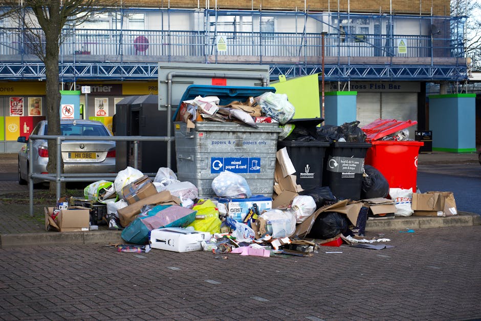 A collection of overflowing waste bins and heaps of household rubbish are situated on a paved urban street near a metal railing. The scene includes a large grey mixed waste bin with the label 'mixed paper & card' that is filled to capacity, causing trash such as cardboard boxes, paper bags, plastic packaging, and packaging materials to spill onto the surrounding area. Adjacent to it are several black and red bins, also filled with refuse, with some bags and loose items on the ground in front. Various discarded items, including flattened cardboard boxes, plastic bottles, paper sheets, and packaging debris, are scattered across the pavement. In the background, there's a building with a blue scaffolding or fencing, and a storefront with signage indicating food or fish bar services. A parked metallic grey car is visible behind the rubbish, close to a green sign attached to the metal railing. The scene highlights typical urban waste accumulation that could be addressed by private rubbish removal services like those offered by Rubbish Removal Acton, especially for on-site clearance or alternative waste handling outside standard municipal collections.