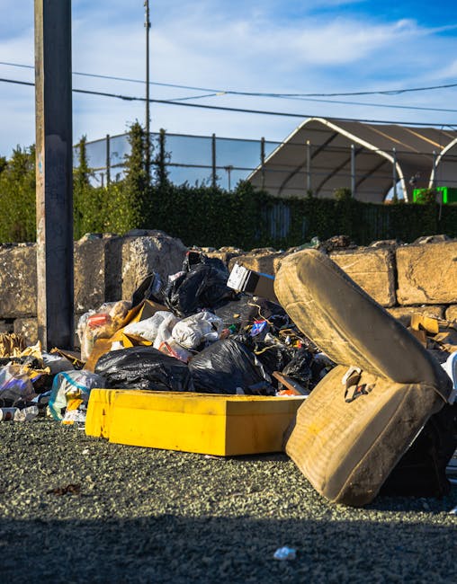A collection of overflowing waste bins and heaps of household rubbish are situated on a paved urban street near a metal railing. The scene includes a large grey mixed waste bin with the label 'mixed paper & card' that is filled to capacity, causing trash such as cardboard boxes, paper bags, plastic packaging, and packaging materials to spill onto the surrounding area. Adjacent to it are several black and red bins, also filled with refuse, with some bags and loose items on the ground in front. Various discarded items, including flattened cardboard boxes, plastic bottles, paper sheets, and packaging debris, are scattered across the pavement. In the background, there's a building with a blue scaffolding or fencing, and a storefront with signage indicating food or fish bar services. A parked metallic grey car is visible behind the rubbish, close to a green sign attached to the metal railing. The scene highlights typical urban waste accumulation that could be addressed by private rubbish removal services like those offered by Rubbish Removal Acton, especially for on-site clearance or alternative waste handling outside standard municipal collections.